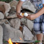 Ein Junge schlägt ein Ei in die Pfanne auf dem Zwergenfeuer