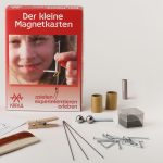 Zwischen dem Experimentierinhalt , z.B. Eisennadeln, Nägel, dem Magnet, Eisenkugeln und Eisenspänen, steht die Schachtel des Kleinen Magnetkastens